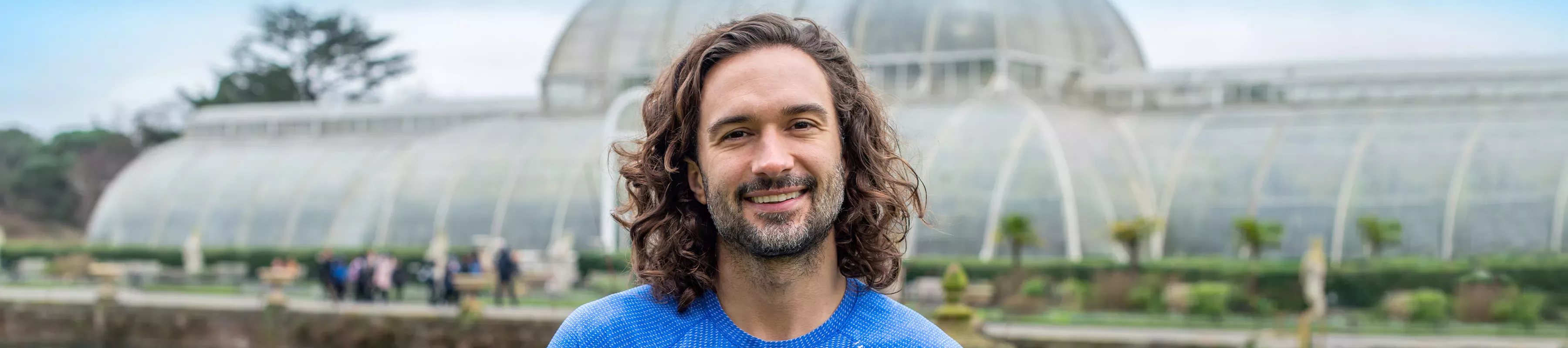 Joe Wicks Festival © RBG Kew A man with long dark hair, Joe Wicks, stood in front of Kews palm House a large glass building.
