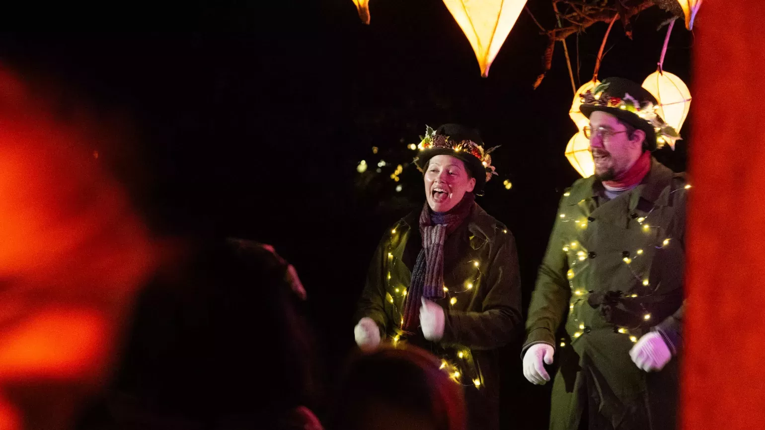 Glow Wild 2023 singers by Jim Holden © RBG Kew Two choral singers in old fashioned trench coats and bowler hats, festooned with fairy lights