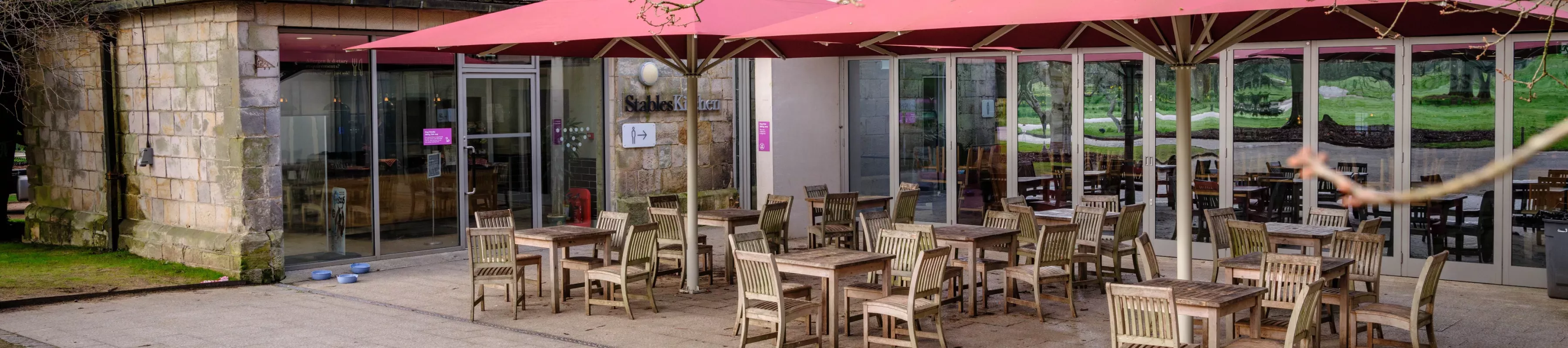 Stables Kitchen © RBG Kew Wooden tables and chairs under red umbrellas outside Wakehurst's Stables Kitchen