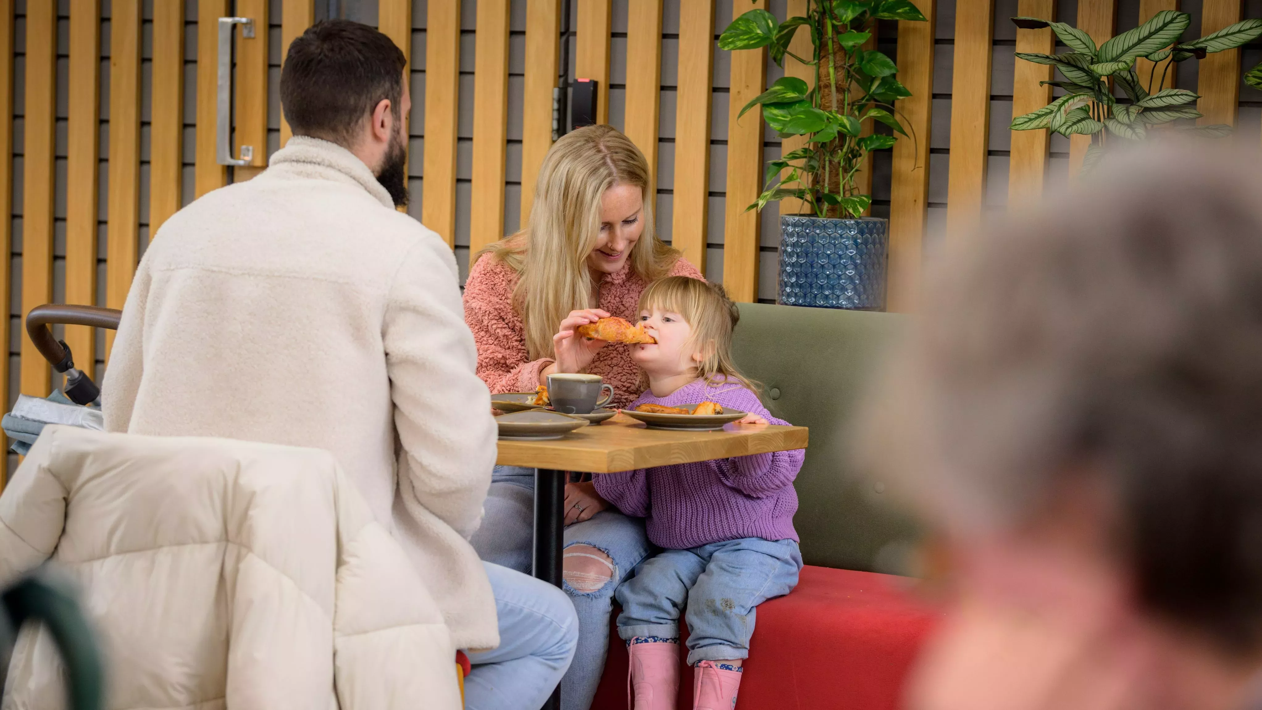 Wakehurst seeds cafe 2023 © RBG Kew A mother feeds a pastry to her young daughter in a cosy cafe while dad looks on