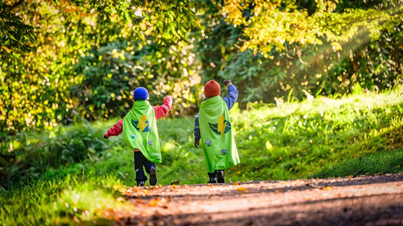 Wakehurst Nature Heroes, Jim Holden © RBG Kew Seen from behind, two children run down a sunny woodland path in autumn, wearing red and blue beanies and green Nature Heroes capes