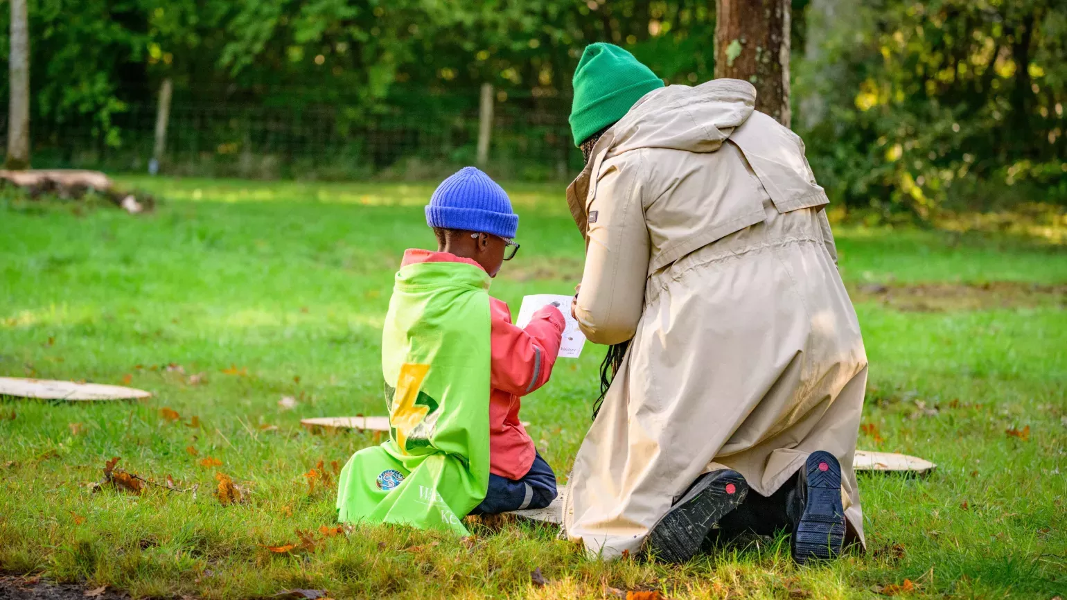 © RBG Kew Seen from behind, a mother kneels next to her child on a grassy lawn at the start of a trail of stepping stones, both of them looking at a sheet of paper