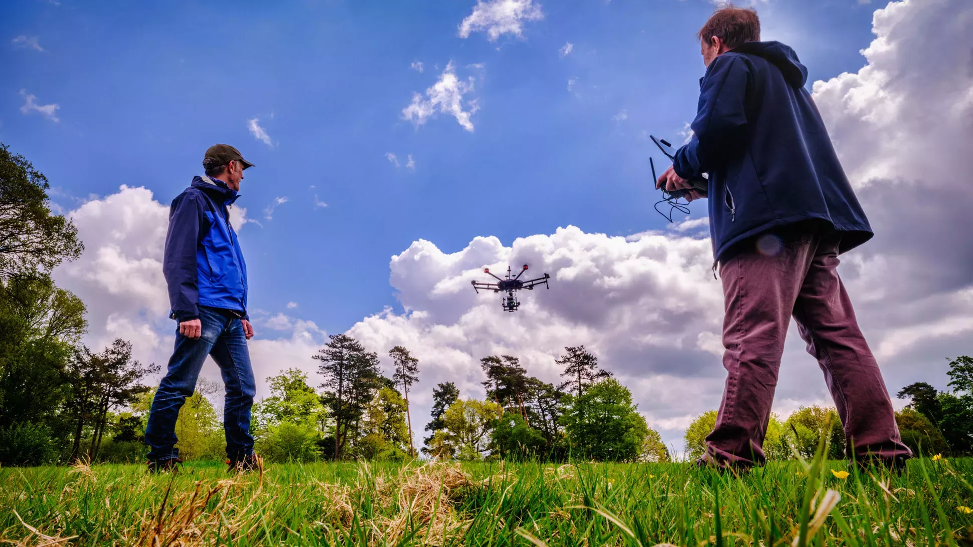 Drones at Wakehurst © RBG Kew. Blue sky above two people in a field watching a drone go up