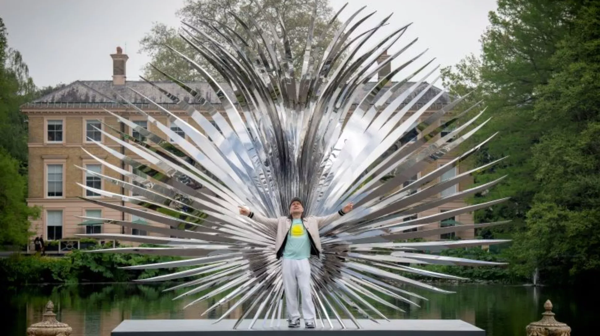 Marc Quinn at Kew, Jeff Eden © RBG Kew Marc Quinn standing in front of a large shiny metal palm leaf sculpture at Kew Gardens