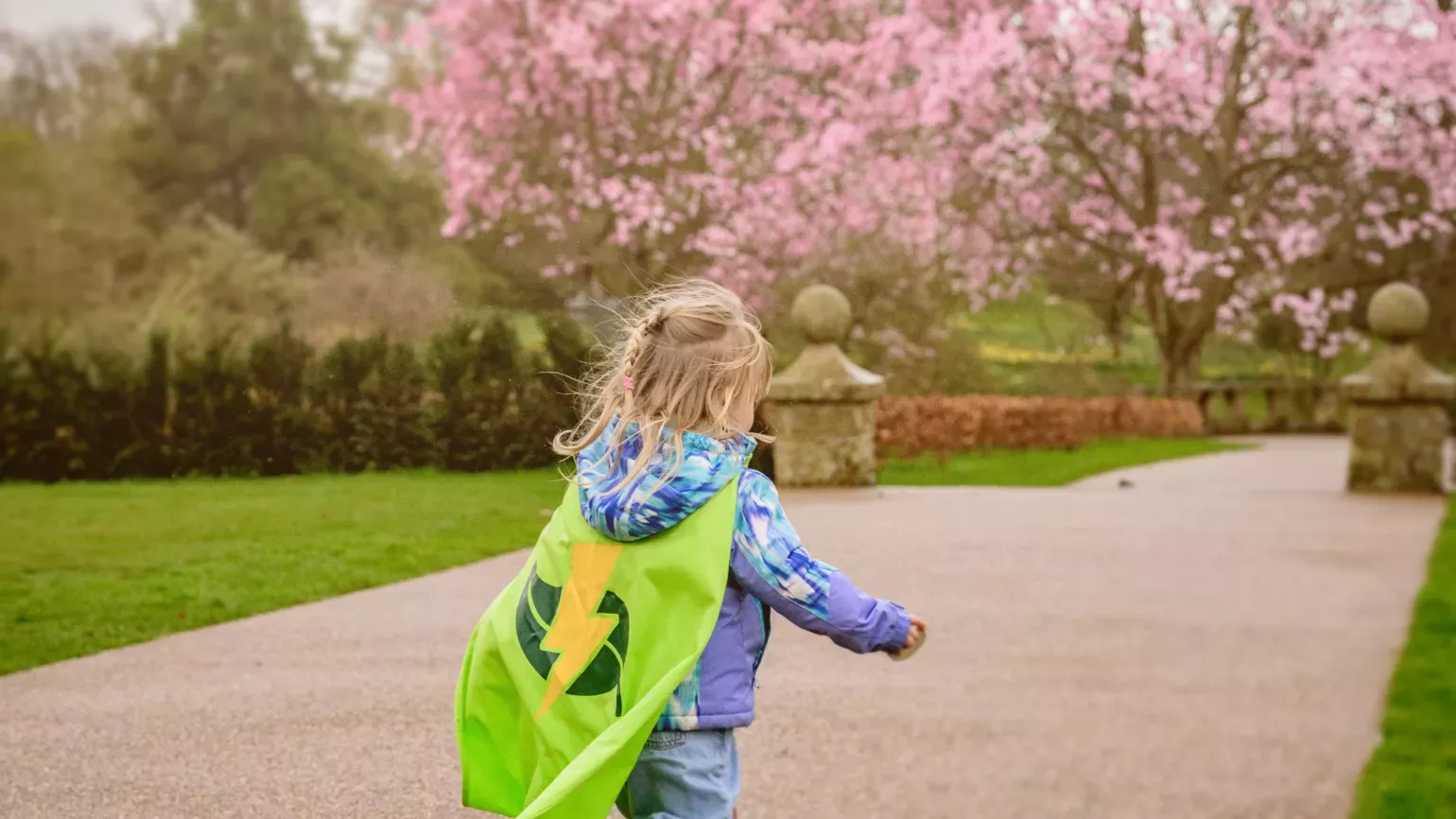 © RBG Kew A small girl in a parka, wellies and green Nature Heroes cape runs down a garden path towards cherry trees in bloom
